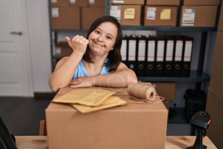 Photo for Hispanic girl with down syndrome working at small business ecommerce pointing thumb up to the side smiling happy with open mouth - Royalty Free Image