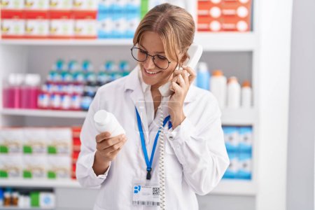 Téléchargez les photos : Young blonde girl pharmacist holding pills bottle talking on telephone at pharmacy - en image libre de droit