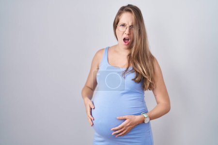 Téléchargez les photos : Jeune femme enceinte debout sur fond blanc dans le visage de choc, l'air sceptique et sarcastique, surpris avec la bouche ouverte - en image libre de droit