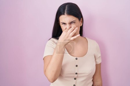 Photo for Young hispanic woman standing over pink background smelling something stinky and disgusting, intolerable smell, holding breath with fingers on nose. bad smell - Royalty Free Image