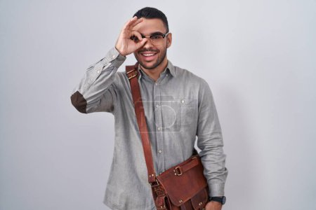 Photo for Young hispanic man wearing suitcase doing ok gesture with hand smiling, eye looking through fingers with happy face. - Royalty Free Image