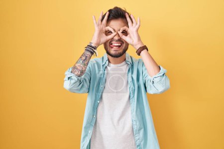 Photo for Young hispanic man with tattoos standing over yellow background doing ok gesture like binoculars sticking tongue out, eyes looking through fingers. crazy expression. - Royalty Free Image