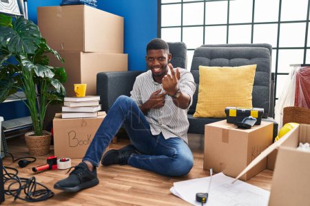 Foto de Hombre afroamericano sentado en el suelo en un nuevo hogar haciendo señas ven aquí gesto con la mano invitando a la bienvenida feliz y sonriente - Imagen libre de derechos