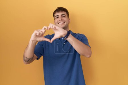 Téléchargez les photos : Jeune homme hispanique debout sur fond jaune souriant dans l'amour faisant forme de symbole de coeur avec les mains. concept romantique. - en image libre de droit