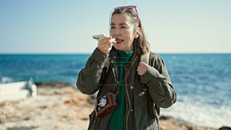 Téléchargez les photos : Femme hispanique mature avec des cheveux gris touriste portant un sac à dos parlant au téléphone au bord de la mer - en image libre de droit