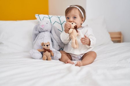 Photo for Adorable caucasian baby sitting on bed bitting doll at bedroom - Royalty Free Image