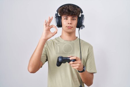 Foto de Adolescente hispano jugando videojuego sosteniendo controlador sonriente positiva haciendo signo ok con la mano y los dedos. expresión exitosa. - Imagen libre de derechos