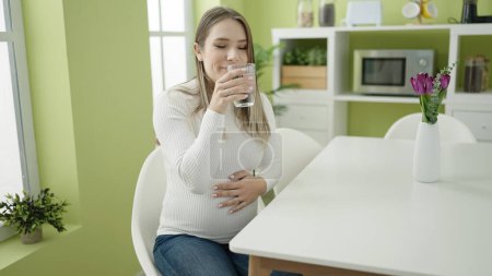 Foto de Joven embarazada bebiendo vaso de agua sentada en la mesa en el comedor - Imagen libre de derechos