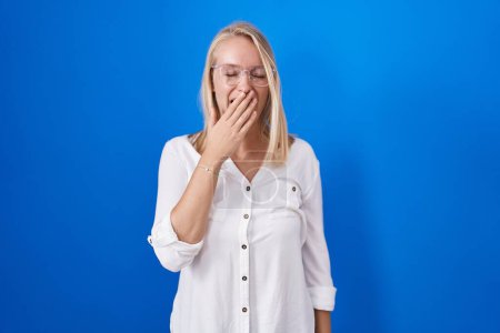 Foto de Young caucasian woman standing over blue background bored yawning tired covering mouth with hand. restless and sleepiness. - Imagen libre de derechos