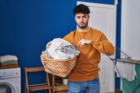 Photo for Hispanic man with beard holding laundry basket at laundry room with angry face, negative sign showing dislike with thumbs down, rejection concept - Royalty Free Image