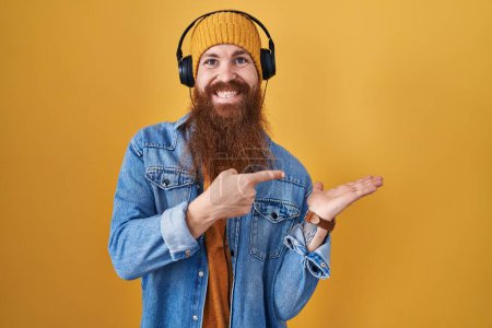 Foto de Hombre caucásico con barba larga escuchando música usando auriculares asombrados y sonriendo a la cámara mientras se presenta con la mano y señalando con el dedo. - Imagen libre de derechos