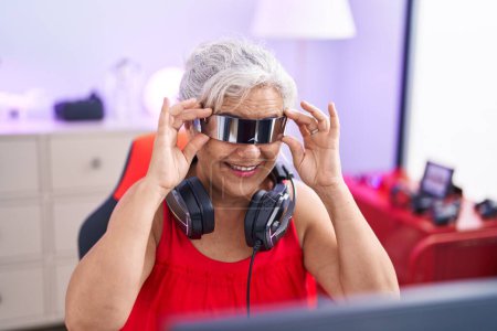 Téléchargez les photos : Femme aux cheveux gris moyen âge streamer jouer à un jeu vidéo en utilisant des lunettes de réalité virtuelle à la salle de jeux - en image libre de droit