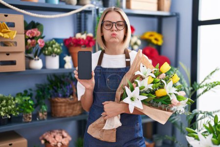 Téléchargez les photos : Jeune femme caucasienne travaillant à la boutique de fleuristes montrant écran smartphone joues gonflées avec drôle de visage. bouche gonflée d'air, attrapant l'air. - en image libre de droit