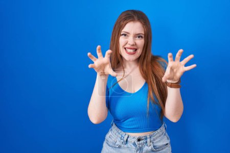 Photo for Redhead woman standing over blue background smiling funny doing claw gesture as cat, aggressive and sexy expression - Royalty Free Image