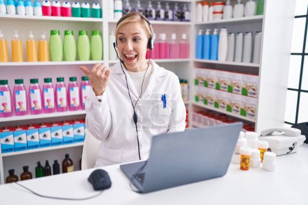 Téléchargez les photos : Jeune femme caucasienne travaillant à la pharmacie pharmacie en utilisant un ordinateur portable souriant avec le visage heureux regardant et pointant vers le côté avec le pouce vers le haut. - en image libre de droit