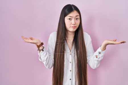 Foto de Mujer joven china de pie sobre fondo rosa expresión despistada y confusa con los brazos y las manos levantadas. concepto de duda. - Imagen libre de derechos
