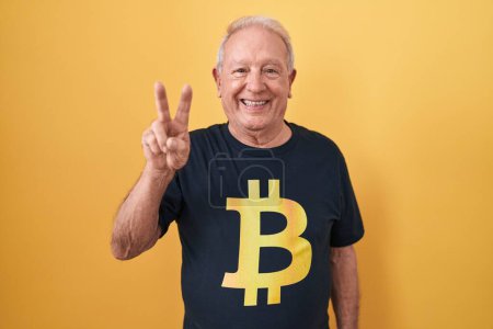 Téléchargez les photos : Homme âgé avec les cheveux gris portant Bitcoin t-shirt souriant avec le visage heureux clin d'oeil à la caméra faisant signe de victoire avec les doigts. numéro deux. - en image libre de droit