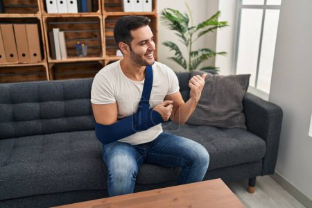 Photo for Young hispanic man with beard wearing arm on sling sitting at therapy consult pointing thumb up to the side smiling happy with open mouth - Royalty Free Image