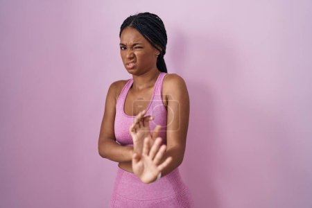 Foto de Mujer afroamericana con trenzas que usan ropa deportiva sobre fondo rosa expresión disgustada, disgustada y temerosa haciendo cara de disgusto porque reacción de aversión. - Imagen libre de derechos