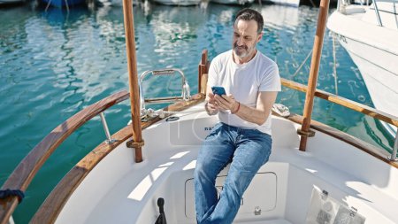 Foto de Hombre de mediana edad con teléfono inteligente sentado en el barco en el puerto - Imagen libre de derechos