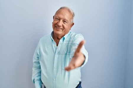 Foto de Hombre mayor con el pelo gris de pie sobre fondo azul sonriendo amistoso ofreciendo apretón de manos como saludo y bienvenida. negocios exitosos. - Imagen libre de derechos