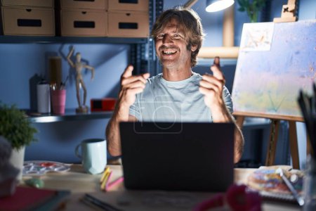 Foto de Hombre de mediana edad sentado en el estudio de arte con portátil por la noche señalando los dedos a la cámara con la cara feliz y divertida. buena energía y vibraciones. - Imagen libre de derechos