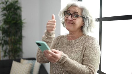 Foto de Mujer de mediana edad con cabello gris usando smartphone con pulgar en casa - Imagen libre de derechos