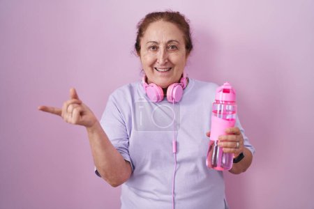 Foto de Mujer mayor con ropa deportiva y auriculares con una gran sonrisa en la cara, apuntando con el dedo de la mano hacia un lado mirando a la cámara. - Imagen libre de derechos