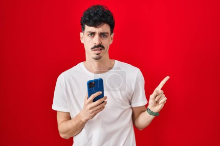 Foto de Hombre hispano usando teléfono inteligente sobre fondo rojo señalando a un lado preocupado y nervioso con el dedo índice, preocupado y expresión sorprendida - Imagen libre de derechos