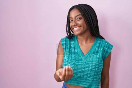 Foto de Joven afroamericano con trenzas de pie sobre fondo rosa señas vienen aquí gesto con la mano invitando a la bienvenida feliz y sonriente - Imagen libre de derechos
