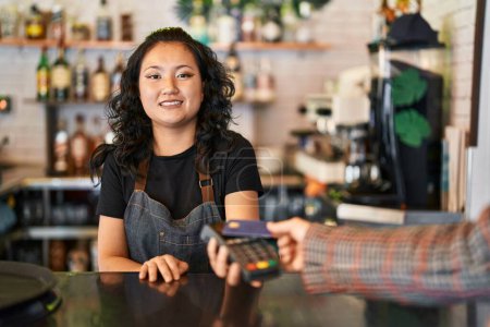 Foto de Joven camarera china sonriendo confiada usando tarjeta de crédito y teléfono de datos en el restaurante - Imagen libre de derechos