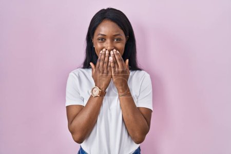 Téléchargez les photos : Jeune femme africaine portant casual t-shirt blanc riant et embarrassé rire couvrant la bouche avec les mains, commérages et concept scandale - en image libre de droit