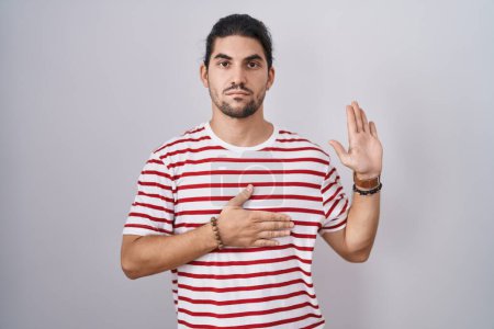 Photo for Hispanic man with long hair standing over isolated background swearing with hand on chest and open palm, making a loyalty promise oath - Royalty Free Image
