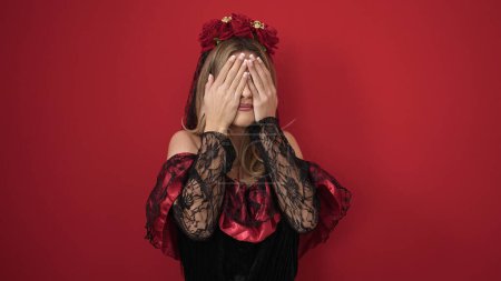 Foto de Mujer rubia joven con traje de katrina cubriendo los ojos con las manos sobre el fondo rojo aislado - Imagen libre de derechos