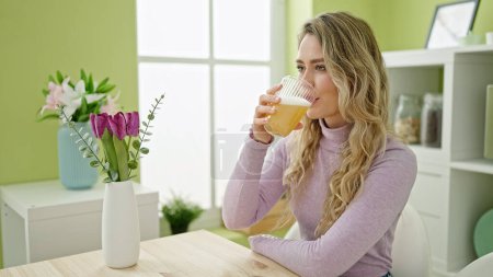 Photo for Young blonde woman drinking glass of orange juice sitting on table at dinning room - Royalty Free Image
