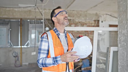 Foto de Constructor hombre de mediana edad cansado usando hardhat como ventilador de mano en el sitio de construcción - Imagen libre de derechos