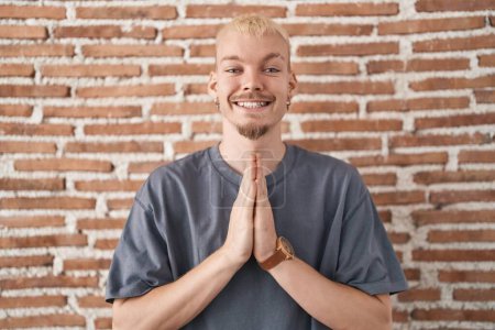 Photo for Young caucasian man standing over bricks wall praying with hands together asking for forgiveness smiling confident. - Royalty Free Image