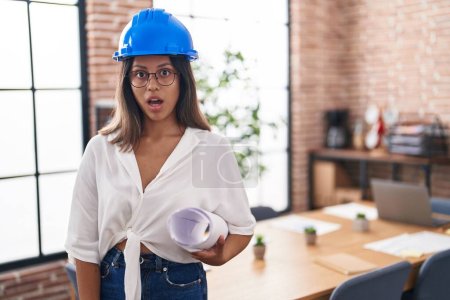 Photo for Hispanic young woman wearing architect hardhat at office scared and amazed with open mouth for surprise, disbelief face - Royalty Free Image