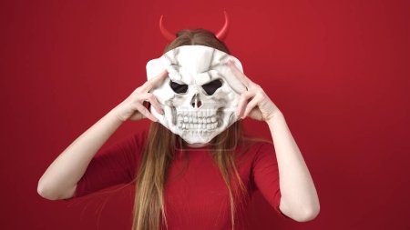 Téléchargez les photos : Jeune femme blonde portant costume diable tenant masque de crâne sur fond rouge isolé - en image libre de droit