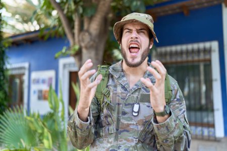 Téléchargez les photos : Jeune homme hispanique portant un uniforme de camouflage de l'armée à l'extérieur fou et fou crier et crier avec une expression agressive et les bras levés. concept de frustration. - en image libre de droit