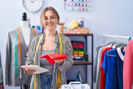 Photo for Young blonde woman tailor smiling confident holding cloths at tailor shop - Royalty Free Image