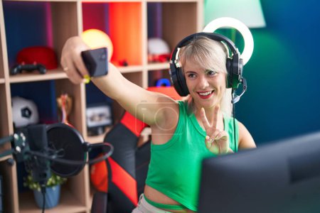Téléchargez les photos : Jeune femme blonde streamer souriant confiant faire selfie par smartphone à la salle de jeux - en image libre de droit