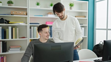 Téléchargez les photos : Deux étudiants hommes utilisant un livre de lecture informatique étudient à l'université de la bibliothèque - en image libre de droit