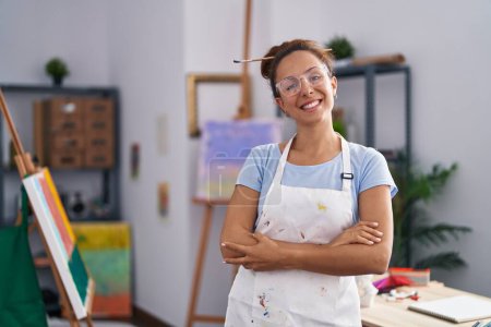 Foto de Mujer morena pintando en el estudio de arte cara feliz sonriendo con los brazos cruzados mirando a la cámara. persona positiva. - Imagen libre de derechos