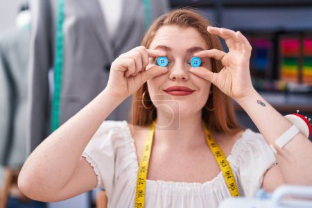 Téléchargez les photos : Jeune femme rousse tailleur souriant confiant tenant des boutons sur les yeux à l'usine de vêtements - en image libre de droit