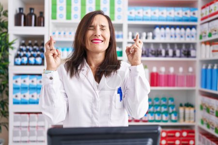Téléchargez les photos : Femme brune d'âge moyen travaillant à la pharmacie drugstore geste doigt croisé sourire avec espoir et les yeux fermés. chance et concept superstitieux. - en image libre de droit