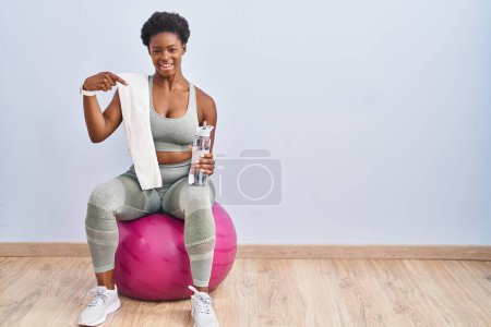 Foto de Mujer afroamericana vestida con ropa deportiva sentada en una pelota de pilates que luce confiada con sonrisa en la cara, señalándose con los dedos orgullosos y felices. - Imagen libre de derechos
