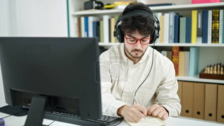 Téléchargez les photos : Jeune homme hispanique étudiant utilisant ordinateur et écouteurs écrivant des notes à l'université de la bibliothèque - en image libre de droit