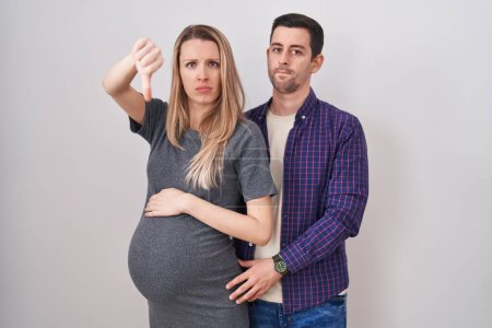 Photo for Young couple expecting a baby standing over white background looking unhappy and angry showing rejection and negative with thumbs down gesture. bad expression. - Royalty Free Image