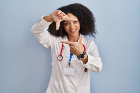 Foto de Joven mujer afroamericana vistiendo uniforme médico y estetoscopio sonriendo haciendo marco con manos y dedos con cara feliz. concepto de creatividad y fotografía. - Imagen libre de derechos